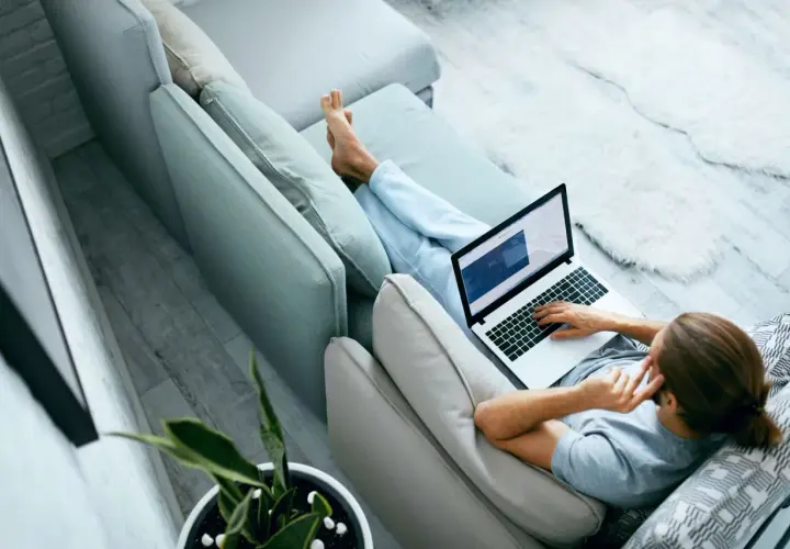 Mężczyzna relaksuje się na kanapie, pracując na laptopie. W tle widać roślinę doniczkową. To idealne miejsce do pracy zdalnej, jak na platformie ozzion.