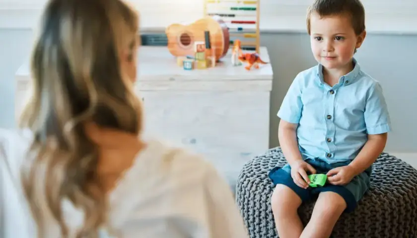 Chłopiec w niebieskiej koszuli siedzi na pufie, rozmawiając z psychoterapeutką.