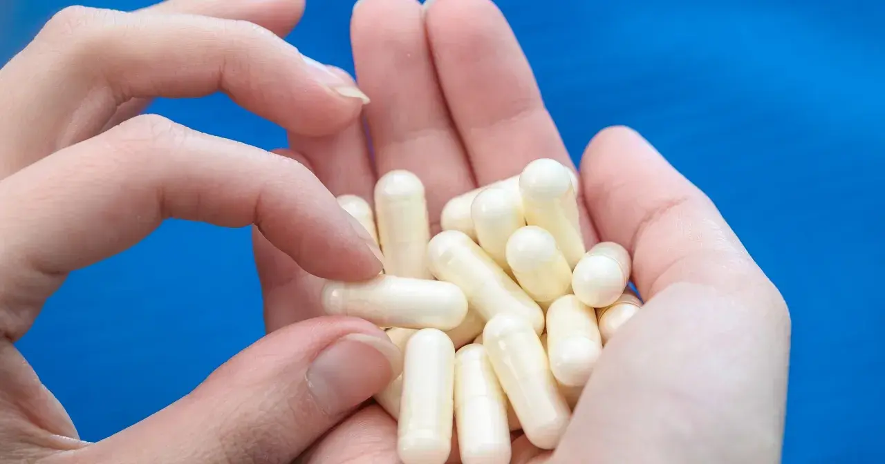 Dłonie trzymają garść białych kapsułek, które mogą zawierać biotynę, wspierającą zdrowie włosów i paznokci.