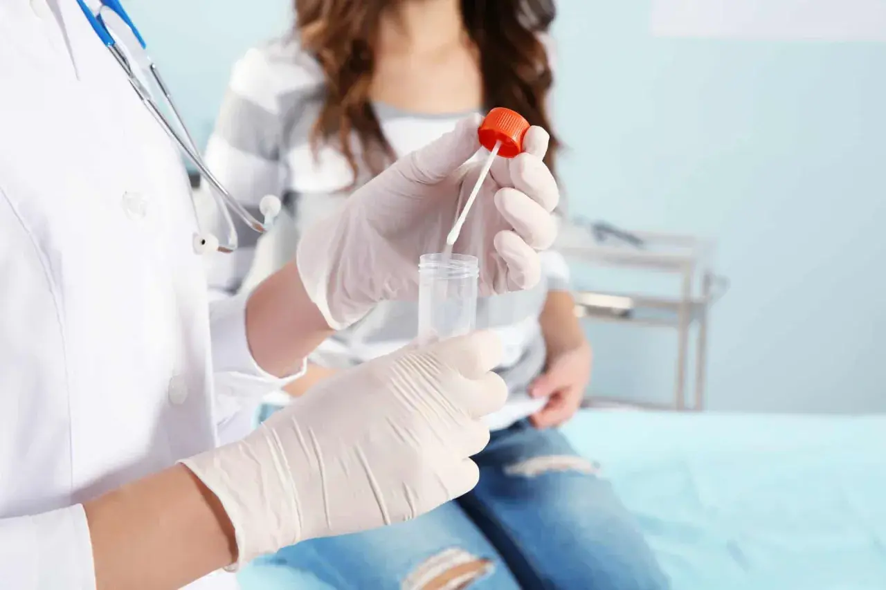 Lekarz w rękawiczkach przygotowuje próbkę do cytologii płynnej. Pacjentka siedzi w tle.